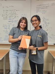 Two women are standing in front of a whiteboard covered in math equations, smiling, and holding an orange book together. They are wearing matching gray t-shirts.