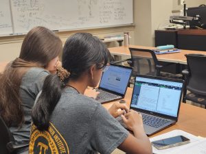 Two individuals are seated at a table, working on laptops in a classroom setting. A whiteboard with equations is in the background. Notebooks and a smartphone are placed on the table beside them.