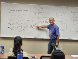 A man stands in front of a whiteboard filled with mathematical equations, pointing to one of them. Several seated individuals face him, paying attention.