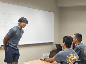 A person standing and speaking in front of a whiteboard with handwritten notes. Three people are seated at a table, attentively listening to the speaker. They all wear gray shirts with a logo.