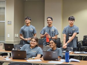 Five students sitting and standing around tables with laptops in a classroom setting, wearing matching gray t-shirts.