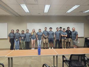 A group of 13 people stand in a classroom, dressed in matching grey shirts. A long table with a blue water bottle and chairs are in the foreground. The room has fluorescent lighting and whiteboards.