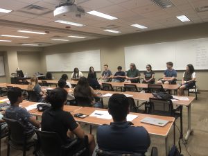 A group of students sits in a panel at the front of a classroom while other students seated at tables listen attentively.