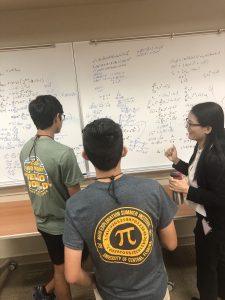 Three people stand in front of a whiteboard filled with complex mathematical equations. Two wear T-shirts with text, while one wears a blazer and holds a cup, appearing to explain something.