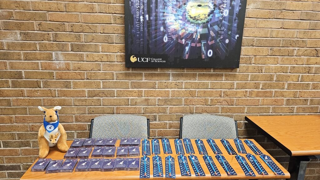 A table with a plush kangaroo, several name tags, and blue lanyards is set up in front of a brick wall and a "UCF College of Sciences" sign.