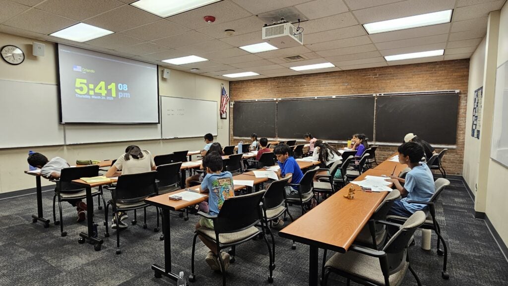 A classroom with students seated at desks working on assignments, a large digital clock on the projector screen reads 5:41 PM, and chalkboards cover the back wall.
