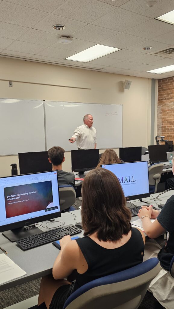 A teacher stands at the front of a classroom speaking to students who are seated at desks with computers, while a presentation is projected on the monitors.