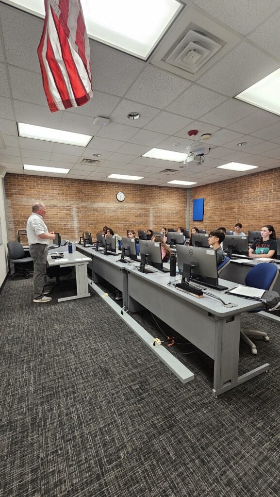 A teacher stands at the front of a computer lab classroom, speaking to students seated at desks with monitors. An American flag hangs from the ceiling.