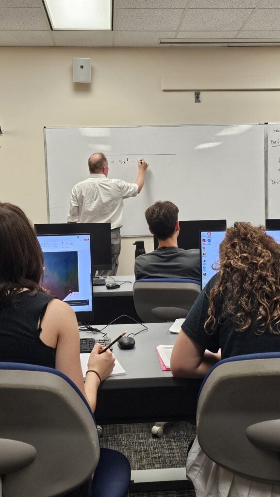 A teacher writes on a whiteboard at the front of a classroom while students sit at desks with computers and take notes.
