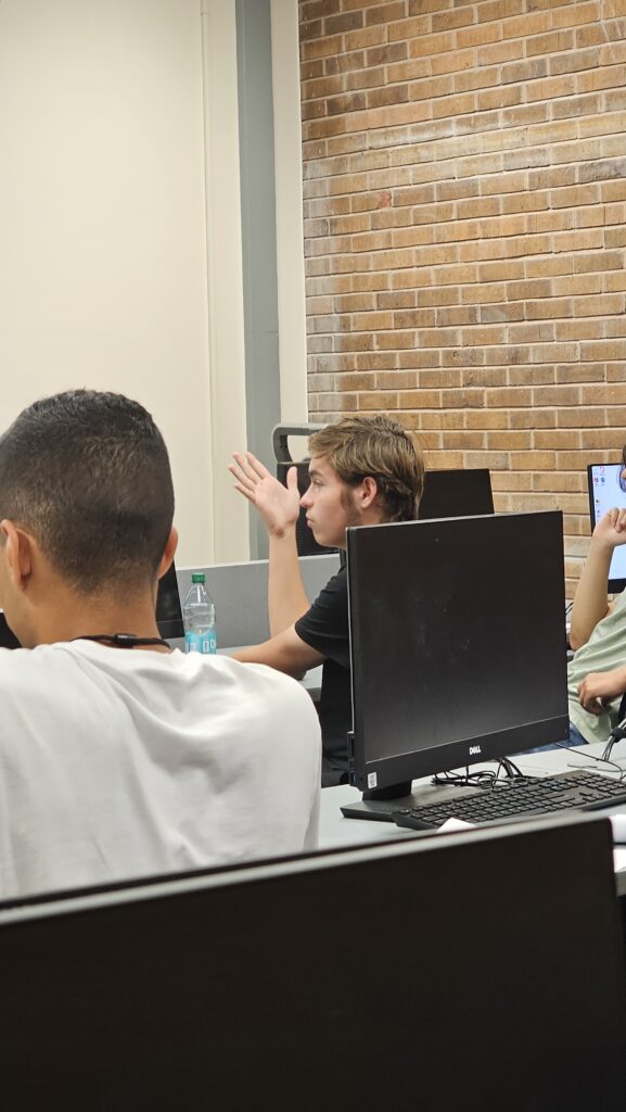 A student gestures with his hand while seated at a computer desk in a classroom; other students and computer monitors are visible.