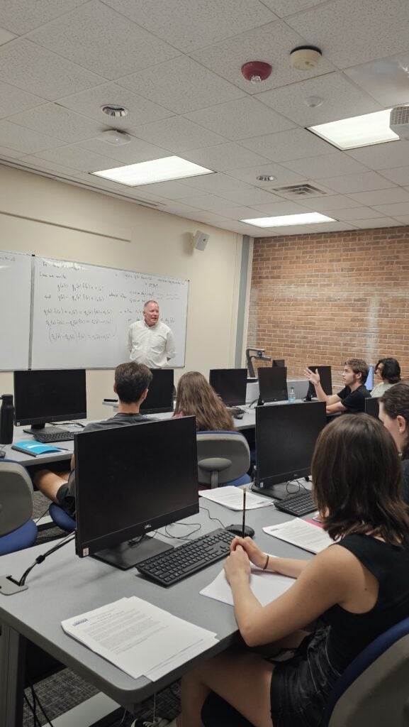 A teacher stands at the front of a classroom speaking to students seated at desks with computers, while notes are written on a whiteboard behind him.