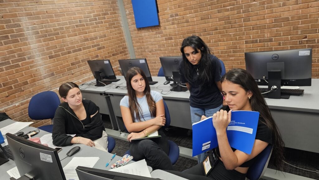 Four women are gathered in an office with computers, discussing documents; one holds a blue folder while the others listen attentively.