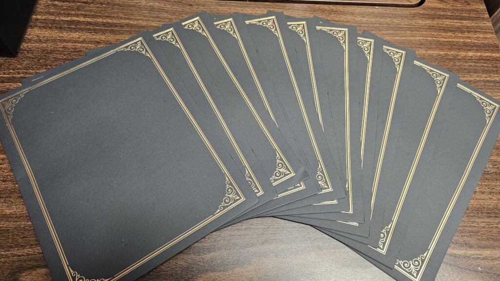 A stack of black paper sheets with ornate gold borders laid out in a fanned arrangement on a wooden surface.