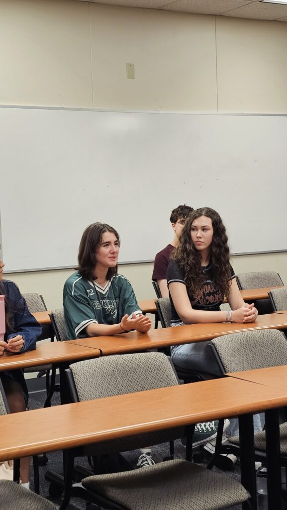 Several students sit in a classroom with empty chairs, facing forward. Two students in the front row appear to be listening or waiting. A large whiteboard is visible behind them.