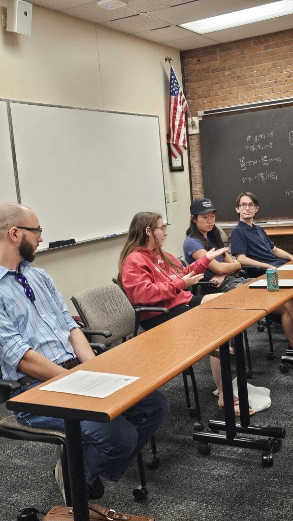 Four people sit at a conference table in a classroom, engaged in discussion. A whiteboard, chalkboard with math equations, and an American flag are visible in the background.