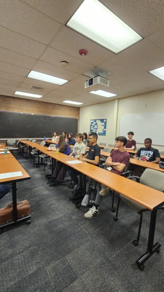A group of students sit at desks in a classroom facing the front, with a chalkboard and projector visible.