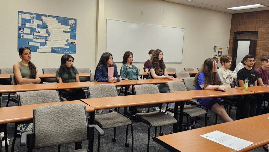 A group of students sits in rows of chairs in a classroom, facing forward, with a whiteboard and bulletin board visible on the walls.
