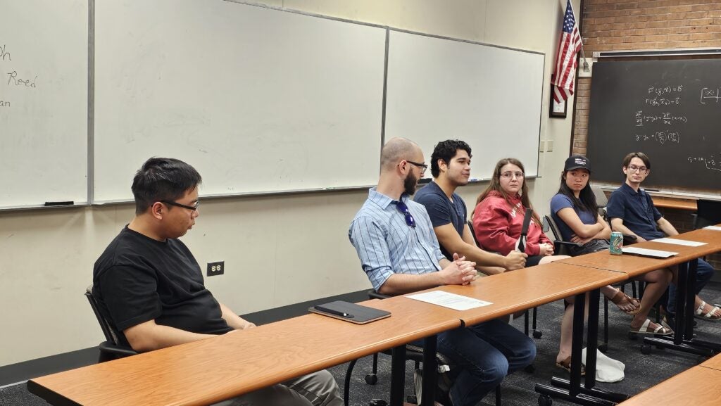 Six people sit at a long table in a classroom, engaged in discussion. A whiteboard, American flag, and notes on the board are visible in the background.