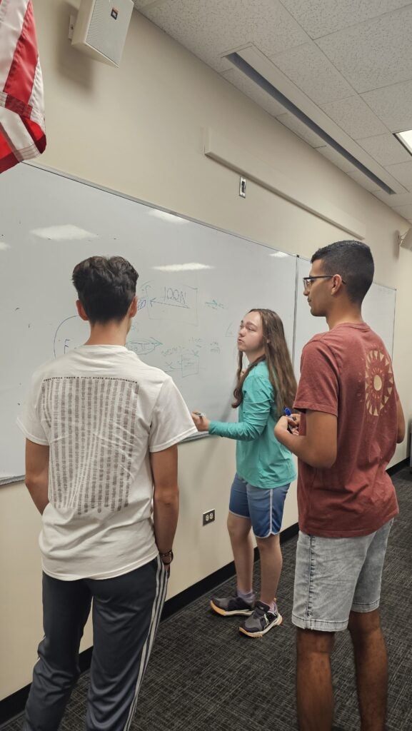 Three young people stand at a whiteboard, with one writing or drawing while the others watch and discuss.