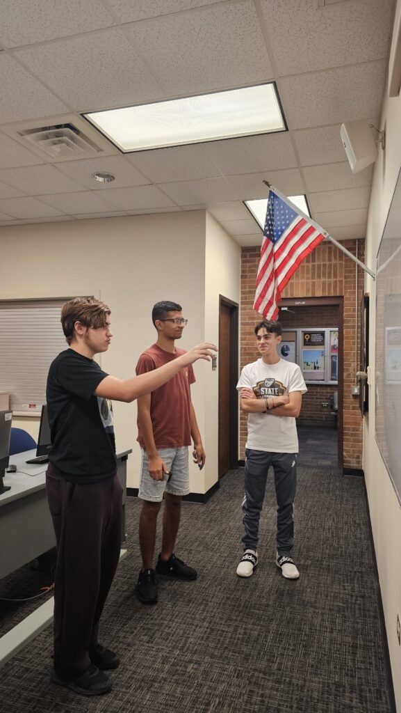 Three young men stand in a classroom near a whiteboard; one gestures towards the board while the others watch. An American flag is mounted on the wall above them.