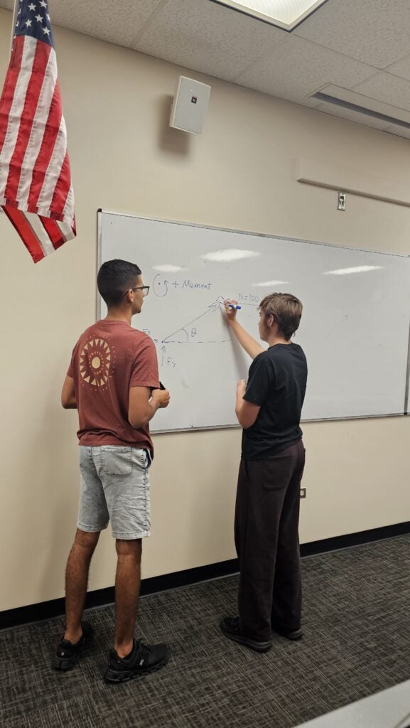 Two students stand at a whiteboard working on a physics diagram, with an American flag hanging on the wall nearby.