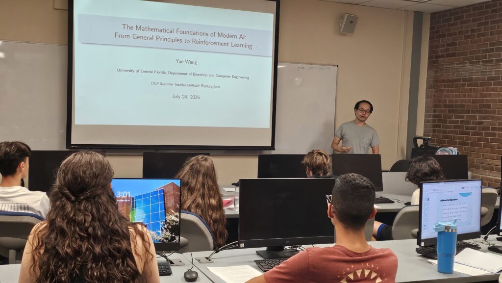 A lecturer stands at the front of a classroom presenting a slide titled “The Mathematical Foundations of Modern AI” to students seated at desks with computers.