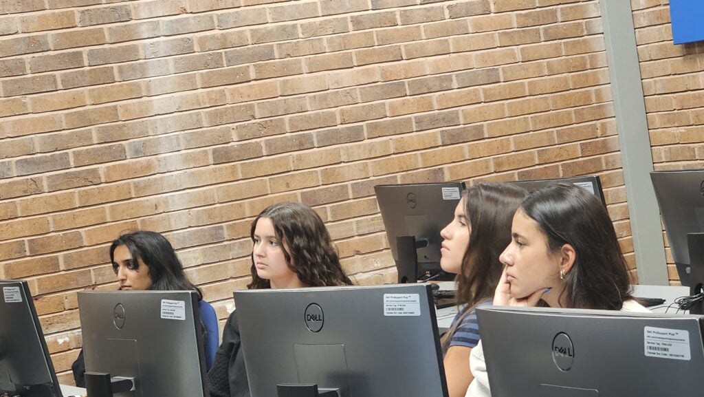 Four students sit at computer desks in a classroom, facing forward and appearing attentive, with a brick wall in the background.