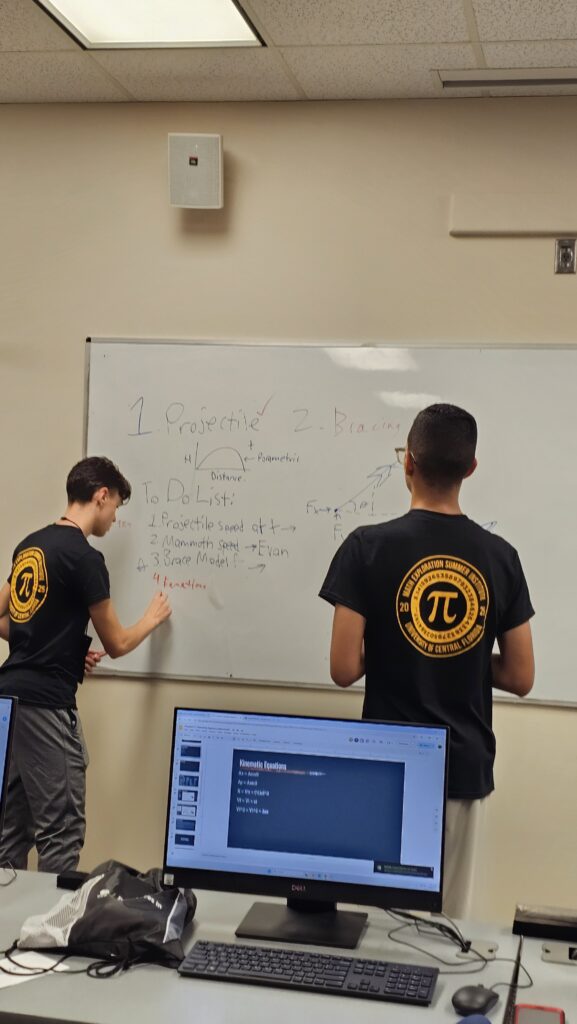 Two students in black Pi-themed shirts write on a whiteboard in a classroom. A computer screen in the foreground displays a list of kinematic equations.