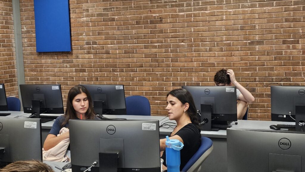 Three students sit at computer desks in a classroom with brick walls. Two are talking, while one in the back appears to be scratching their head.