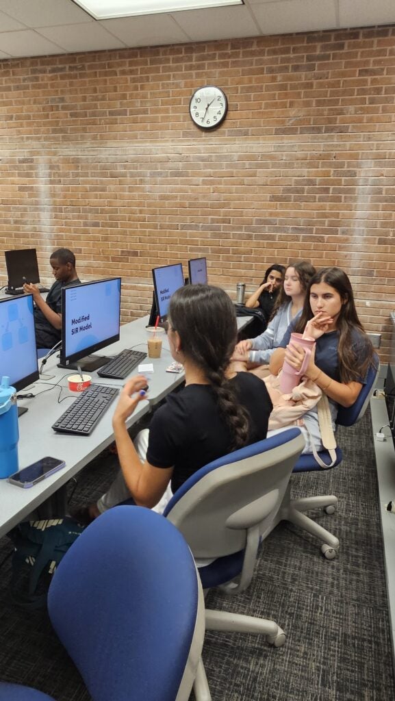 Several students sit at desks using desktop computers in a classroom with a brick wall and a clock. Some have drinks and snacks, and one screen displays "Midfield SRI Media.