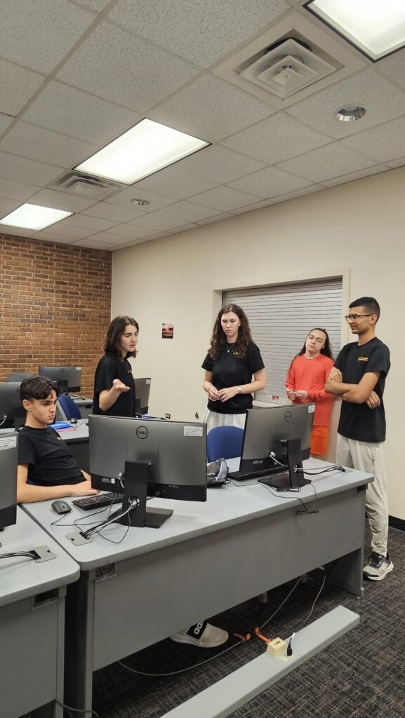 A group of five young adults stand and sit around computer desks in an office or classroom, engaged in discussion.