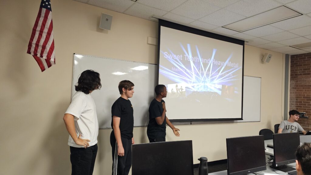 Three students stand at the front of a classroom presenting a slideshow titled "Sweet Toothend/Heavy Metal" while others sit at computers.