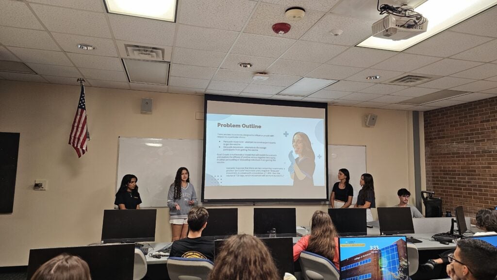 Four people stand at the front of a classroom giving a presentation, with a projected slide titled "Problem Outline" visible behind them. Students sit facing the presenters.