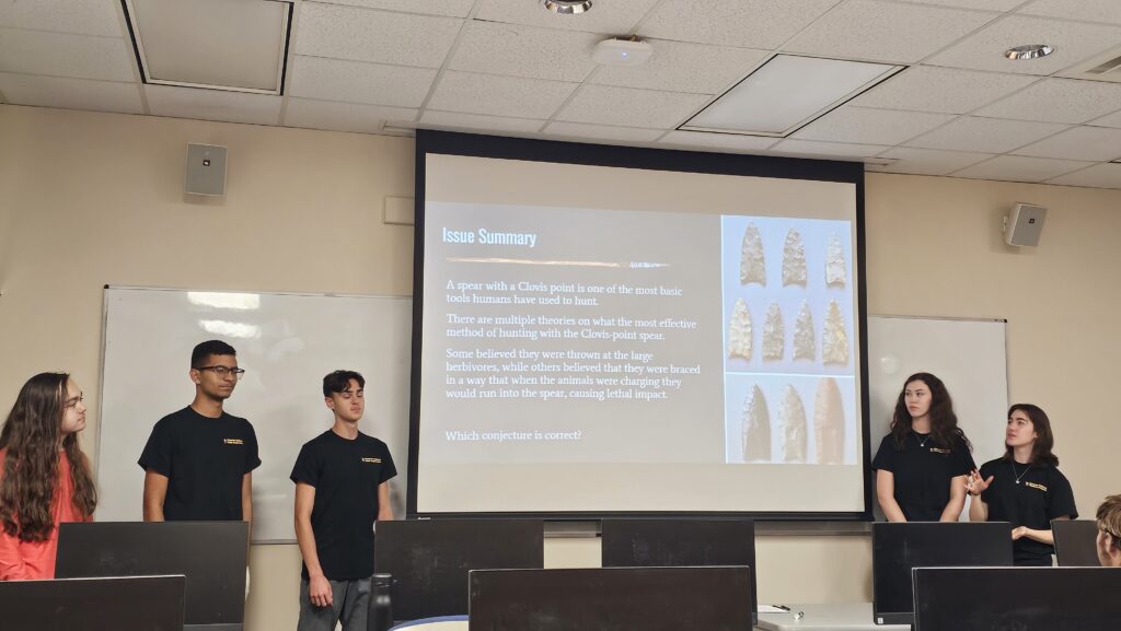 Five students stand at the front of a classroom, presenting a slide about Clovis points and early hunting tools, with images of stone points displayed on the screen behind them.
