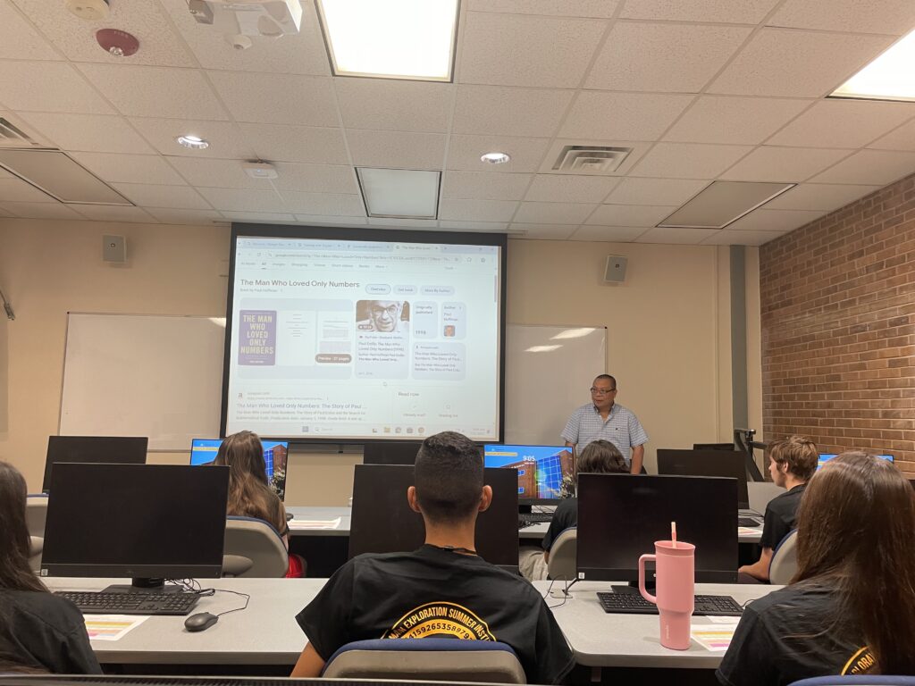A classroom with students seated at computers facing a projector screen, where a man is presenting information about "The Man Who Loved Only Numbers.