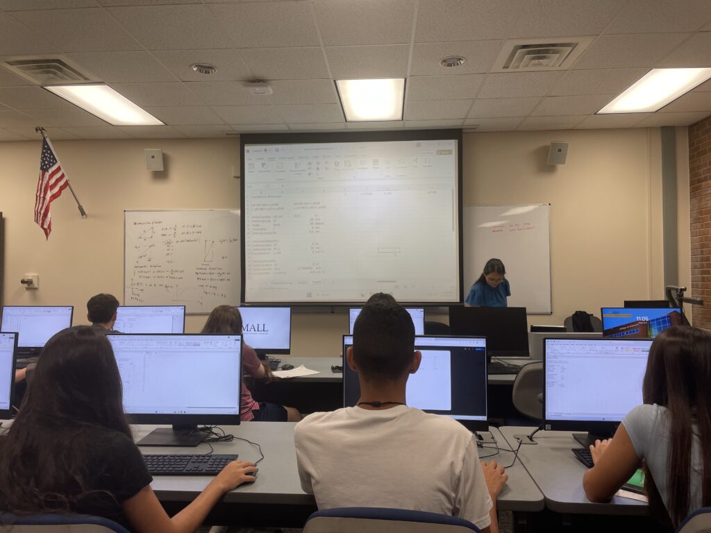 Students sit at computer desks in a classroom, facing a projector screen displaying a spreadsheet, while a teacher stands near a whiteboard at the front.