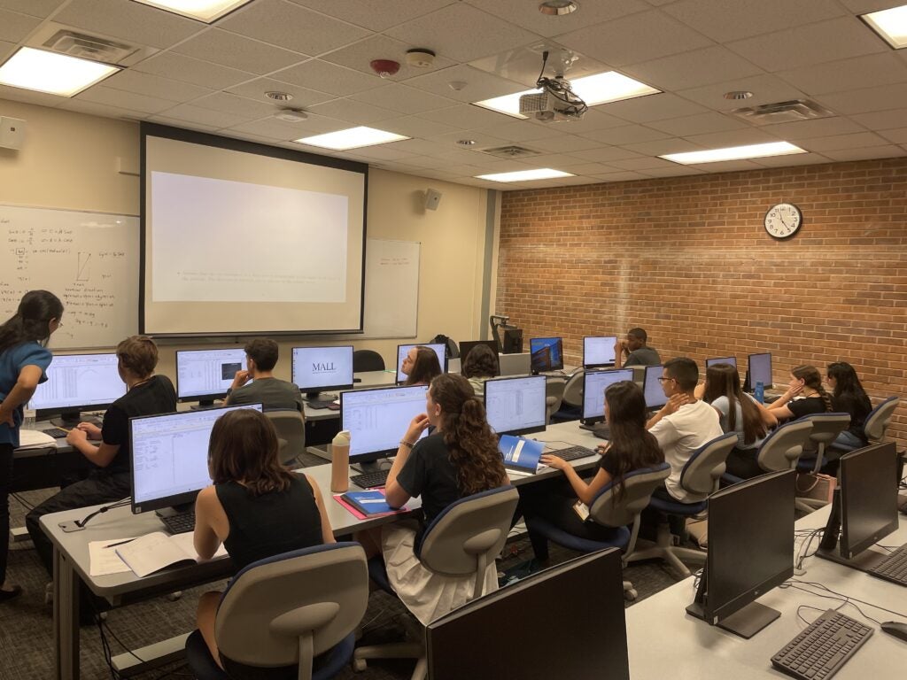 A classroom of students sits at desks with computers, facing a projector screen while an instructor stands at the front explaining content.