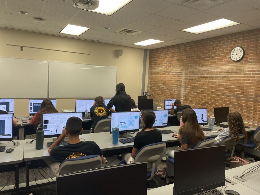 A classroom with students seated at computers facing a whiteboard, while an instructor stands at the front; a clock on the wall shows it is 10:11.