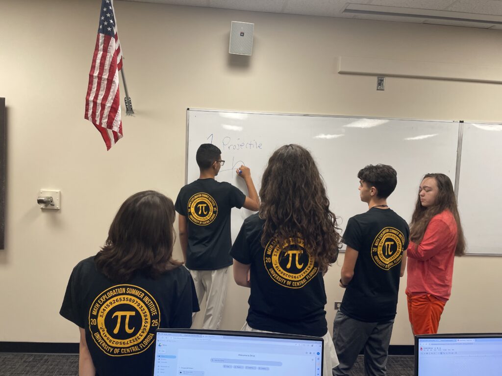 Five students stand at a whiteboard in a classroom, with one writing while others watch. Most are wearing matching black Pi-themed shirts. An American flag hangs on the wall.