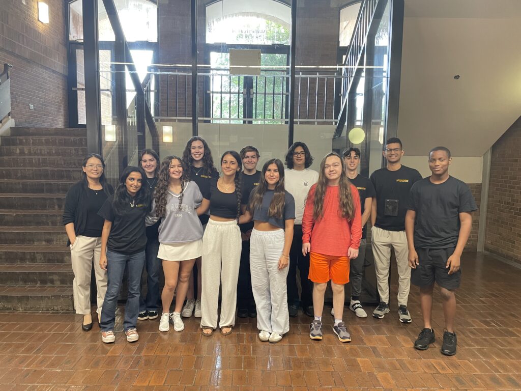 A group of thirteen young people and one adult stand indoors on a brick floor in front of stairs, posing for a group photo.