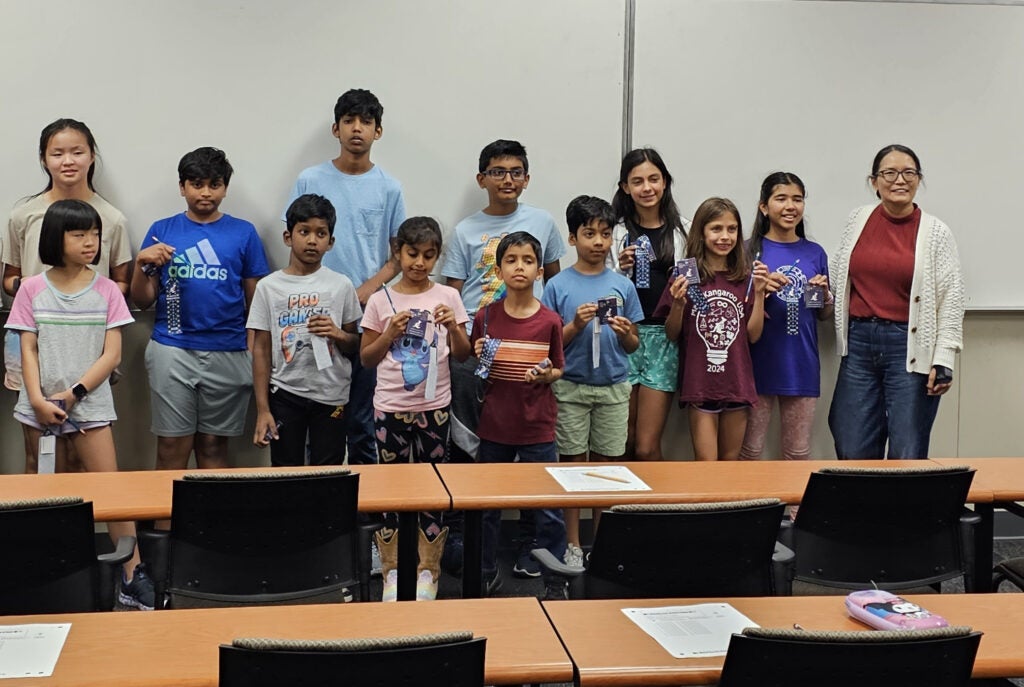 A group of children and one adult stand in a classroom in front of a whiteboard, holding small crafts. Desks with papers and pencil cases are visible in the foreground.