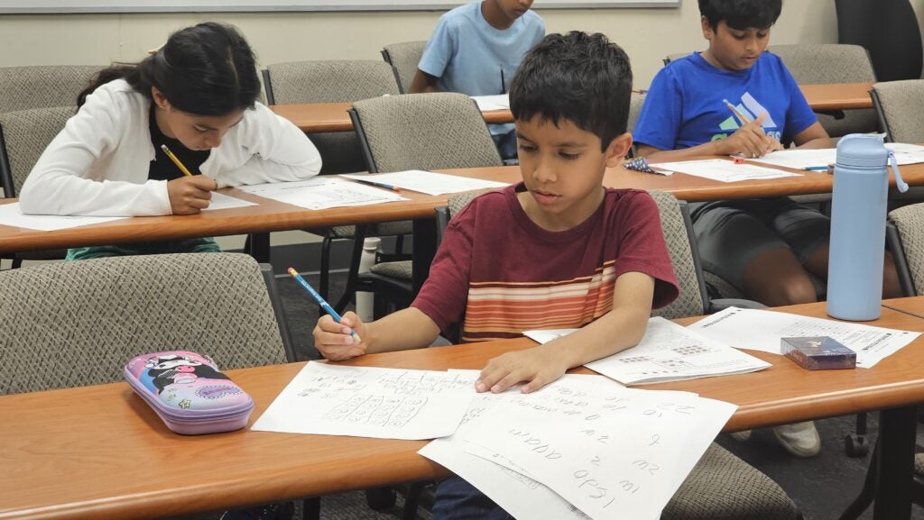 Several children sit at desks in a classroom, working on worksheets with pencils. Papers and stationery are scattered on the desks.
