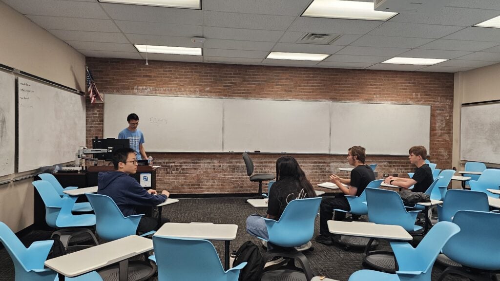 A teacher stands at a podium in front of a whiteboard while five students sit in blue chairs in a classroom.