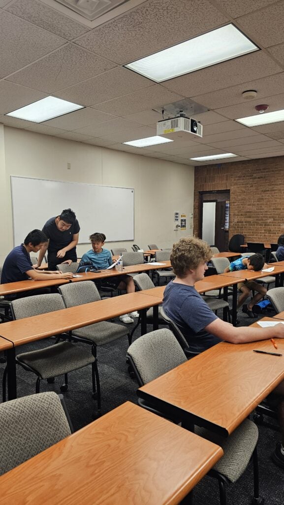 Several students sit and work at desks in a classroom while one person stands, assisting two students. The room has rows of empty chairs and tables, whiteboards, and a ceiling-mounted projector.