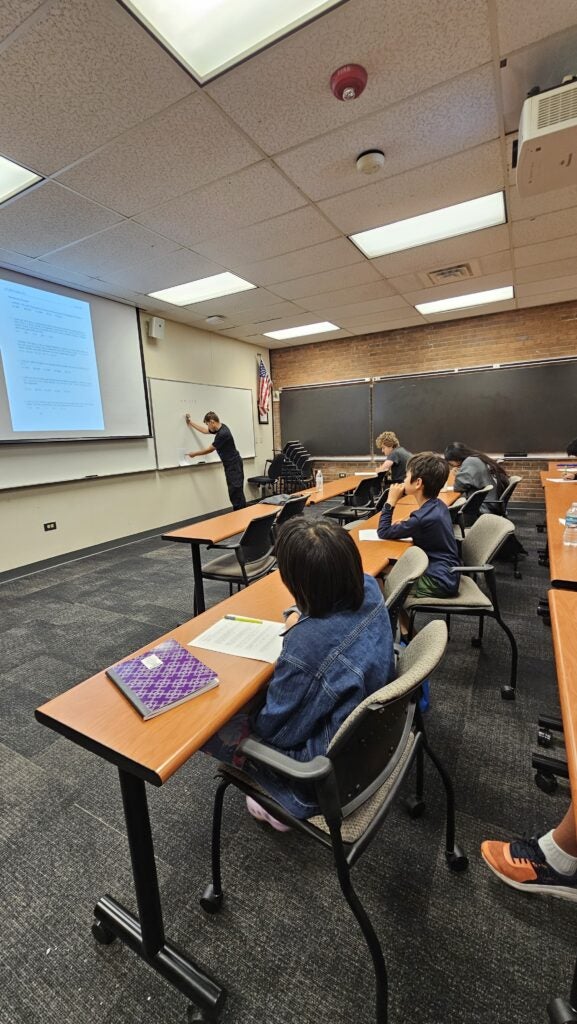 A teacher writes on a whiteboard in a classroom while several students sit at desks, facing the front and taking notes.