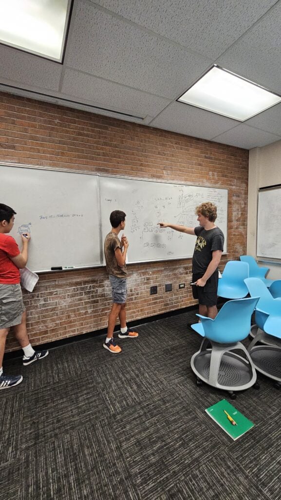 Three students discuss and write on a large whiteboard filled with diagrams and notes in a classroom with blue chairs and a green notebook on the floor.
