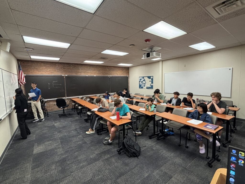 A classroom with students seated at desks, listening to two people standing at the front near blackboards.