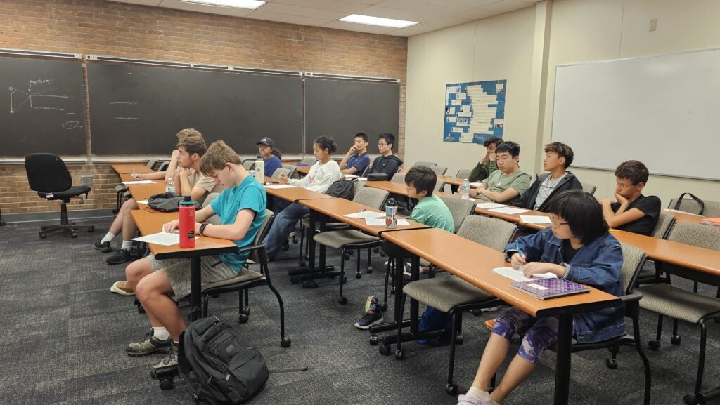 Students sit at desks in a classroom, listening and writing notes. The room has blackboards, a whiteboard, and a bulletin board on the walls.
