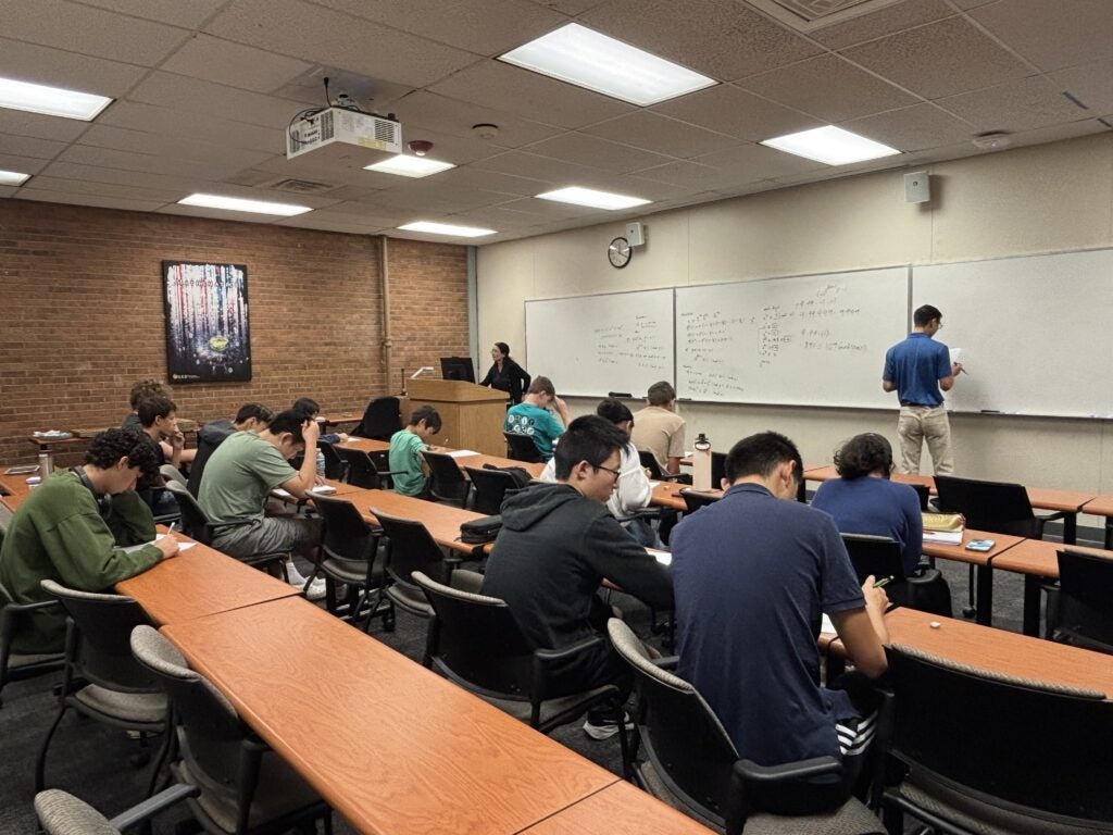 Students sit at desks in a classroom, writing while one person writes on a whiteboard and another stands at a podium.