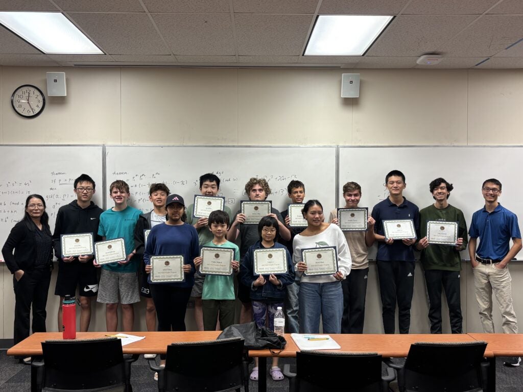 A group of students and two adults stand indoors holding certificates, posing for a photo in front of a whiteboard.